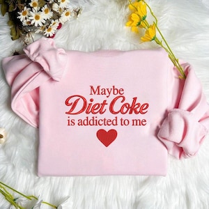 May include: A pale pink sweatshirt featuring the embroidered text "Maybe Diet Coke is addicted to me" in red. A red heart is embroidered below the text. The sweatshirt has long sleeves and a crew neck.