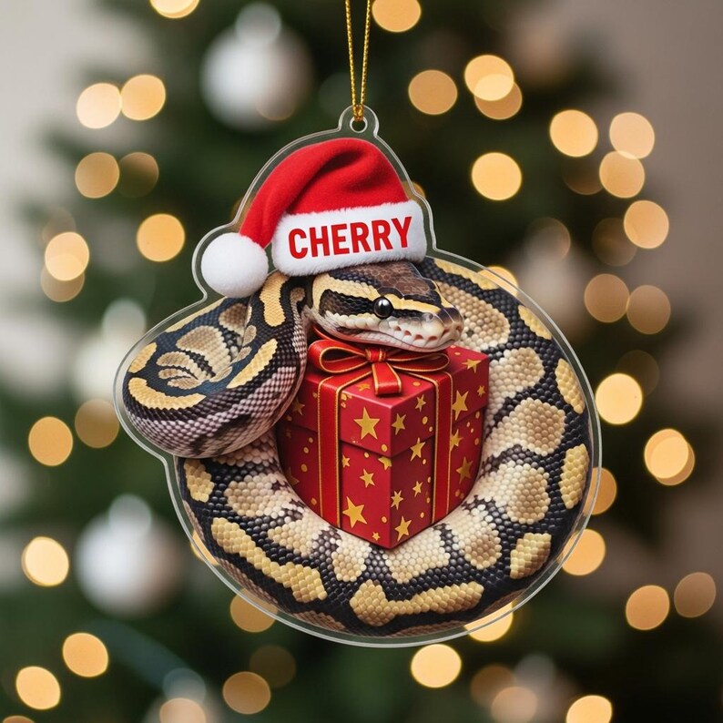 K&ouml;nnte beinhalten: Ein klarer Weihnachtsornament aus Acryl mit einer Schlange, die einen Weihnachtsmannhut mit dem Wort "CHERRY" tr&auml;gt und sich um eine rote Geschenkbox mit goldenen Sternen windet. Die Schlange hat ein braunes und beiges Muster.