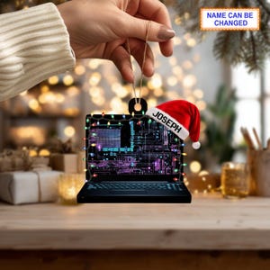 May include: A black laptop-shaped Christmas ornament with a red Santa hat that says "JOSEPH". The ornament is decorated with colorful lights and a circuit board design. The text "NAME CAN BE CHANGED" is also visible.