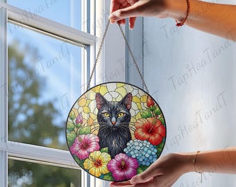 May include: Round stained glass window art featuring a black cat with yellow eyes surrounded by colorful flowers. The artwork is suspended by a chain and is held in front of a window. The flowers include red, pink, yellow, and blue blossoms.