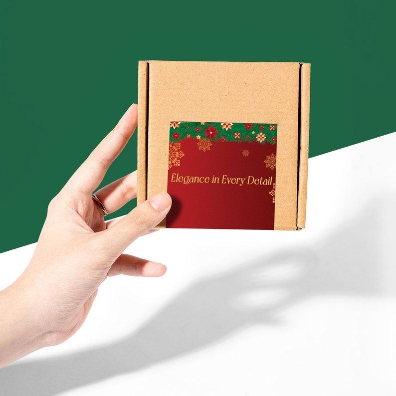 May include: A small, square, brown cardboard box with a red and green label. The label features gold snowflakes and the text "Elegance in Every Detail." The box is held against a green and white background.