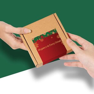 May include: A small, brown cardboard box with a red and green label featuring the text "Elegance in Every Detail." The label has a festive design with snowflakes and floral accents. The box is being held by two hands against a green background.