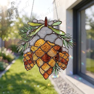 Könnte beinhalten: Ein Buntglas-Sonnenfänger mit einem zentralen Tannenzapfen aus bernsteinfarbenem und braunem Strukturglas, gekrönt von weißem Glas, das Schnee darstellt, und grünen Glas-Tannennadeln. Das Stück ist an zwei dünnen Metallketten aufgehängt.