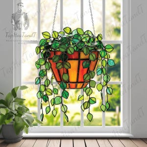 May include: Stained glass hanging planter with a vibrant orange and yellow basket, filled with lush green leaves. The planter is suspended by chains in front of a window. The text "Tap Hoa TandT" is visible.