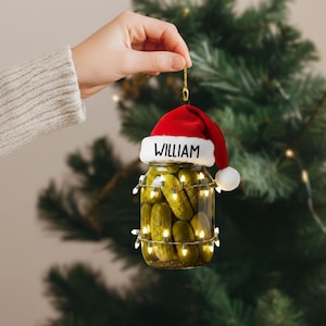May include: A clear glass jar filled with pickles, adorned with a red Santa hat that reads "WILLIAM" in white letters. The jar is wrapped with a string of small, warm white lights, creating a festive Christmas ornament.
