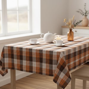 Puede incluir: Un mantel rectangular con un estampado a cuadros en tonos marrones, naranjas y blancos cubre una mesa de madera. Una tetera blanca, dos tazas de té y una vela están sobre la mesa. También hay un jarrón con flores secas.