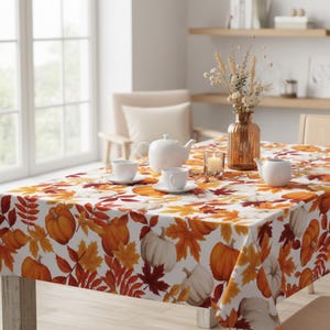 Könnte beinhalten: Ein weißes Tischtuch mit einem herbstlichen Muster aus Kürbissen und Blättern in Orange, Gelb und Rot. Der Tisch ist mit weißen Teetassen, einer Teekanne und einer dekorativen Vase gedeckt. Eine brennende Kerze.