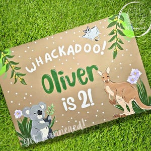 May include: A hand-painted sign on a brown background with the words "WHACKADOO! Oliver is 2!" in white and green. It features illustrations of a kangaroo, koala, and other Australian animals, with floral accents.