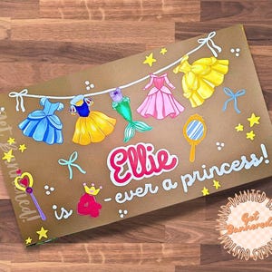 May include: A rectangular sign featuring a brown background with a hand-painted illustration of a clothesline holding various colorful princess dresses, a mirror, and a wand. The text reads, Ellie is 4 - ever a princess! with yellow stars.