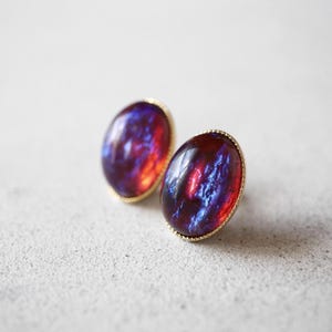 May include: A pair of oval stud earrings featuring dark red and deep blue iridescent stones with a swirling pattern. Each stone is encased in a thin, textured gold-toned metal bezel setting that frames the smooth, rounded surface of the cabochon.