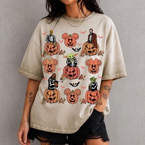 May include: A beige short-sleeve t-shirt featuring a graphic print of various characters wearing skeleton costumes sitting on orange jack-o'-lanterns, interspersed with pumpkin-shaped mouse ears and small orange bats on a light background.