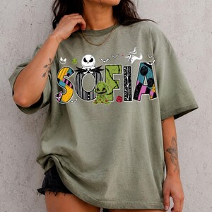 May include: An olive green short-sleeve t-shirt featuring a graphic print of the name SOFIA in stylized letters. The letters incorporate patterns and characters from The Nightmare Before Christmas, including Jack Skellington and Oogie Boogie.