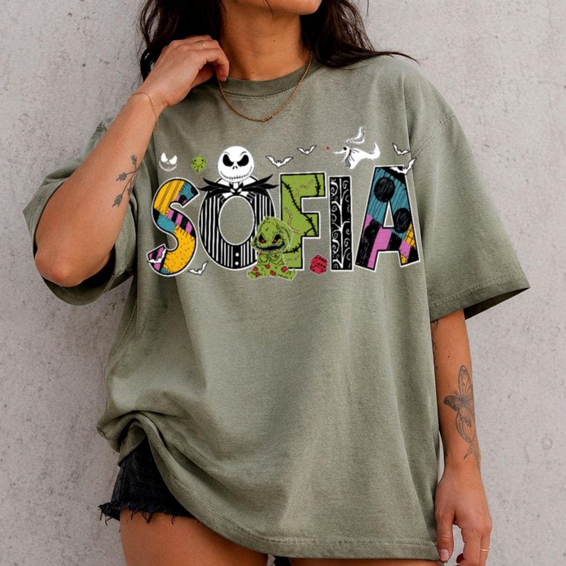 May include: An olive green short-sleeve t-shirt featuring a graphic print of the name SOFIA in stylized letters. The letters incorporate patterns and characters from The Nightmare Before Christmas, including Jack Skellington and Oogie Boogie.