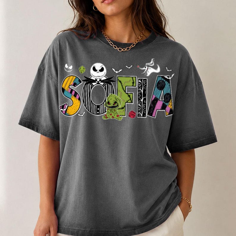 May include: A charcoal gray short-sleeve t-shirt featuring the name SOFIA in large, patterned letters. The design includes a Jack Skellington character, a green Oogie Boogie figure, a ghost dog, bats, and dice graphics in a colorful, patchwork style.