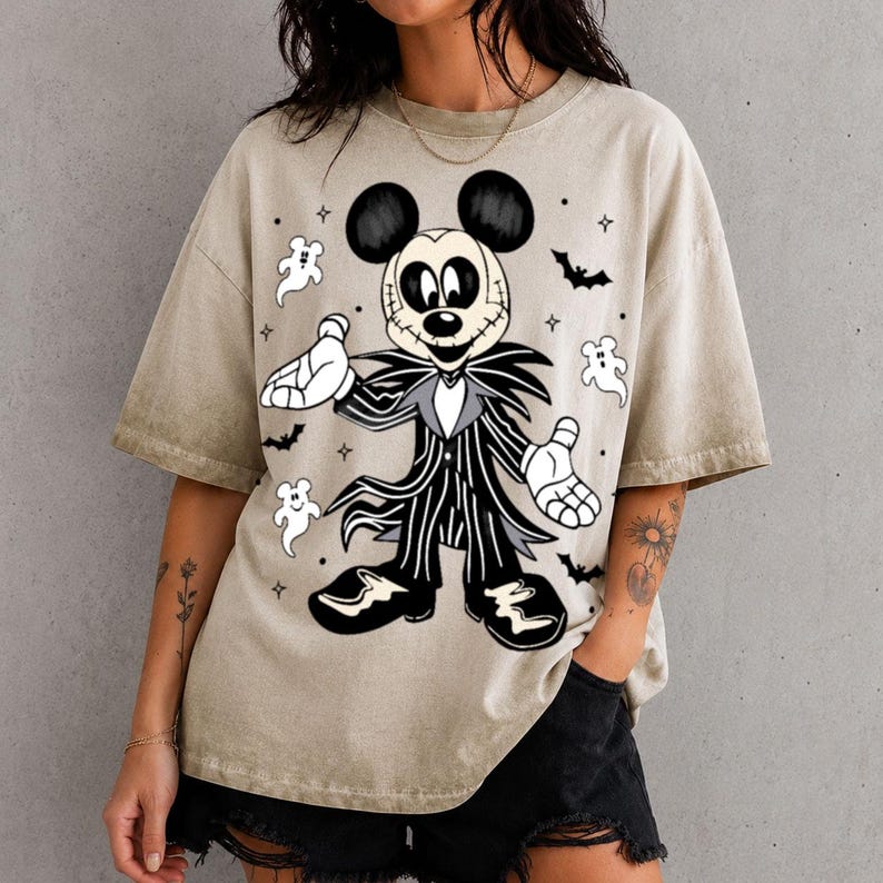 May include: A beige short-sleeve t-shirt featuring a graphic print of a character resembling Mickey Mouse dressed in a black and white pinstriped suit with a stitched face. The design includes small white ghosts, black bats, and stars.