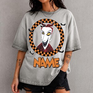 May include: A gray acid-wash t-shirt featuring a graphic of a character with spiky red hair and a wide grin inside a black and orange checkered circle. Small black bats surround the circle, and the word NAME is printed in orange below it.