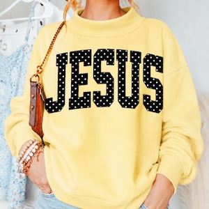 May include: A pale yellow long-sleeve sweatshirt featuring a mock neck collar and the word JESUS printed in large, bold black block letters with a white polka dot pattern across the center of the chest area.