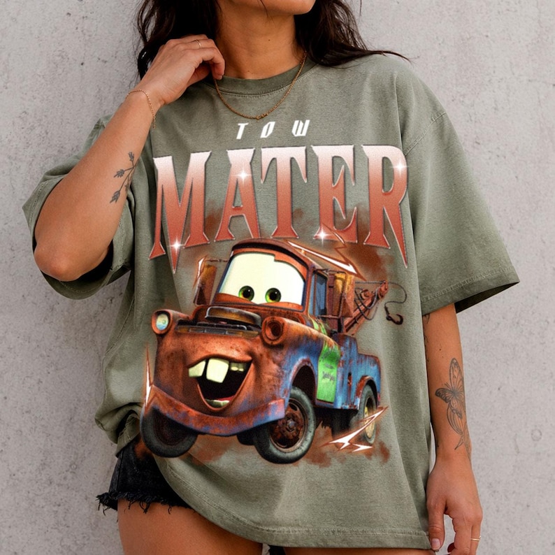 K&ouml;nnte beinhalten: Ein olivgr&uuml;nes Kurzarm-T-Shirt mit einem gro&szlig;en Grafikdruck der Figur Hook aus dem Film Cars. Der Text MATER ist in einer fetten, abgenutzten roten Schrift &uuml;ber der Illustration eines rostigen Abschleppwagens auf das T-Shirt gedruckt.
