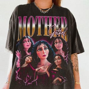 May include: Dark gray t-shirt featuring the text "MOTHER Gothel" above a graphic of the character Gothel from the animated film Tangled. The graphic includes multiple images of Gothel in various poses, with a purple and pink color scheme.