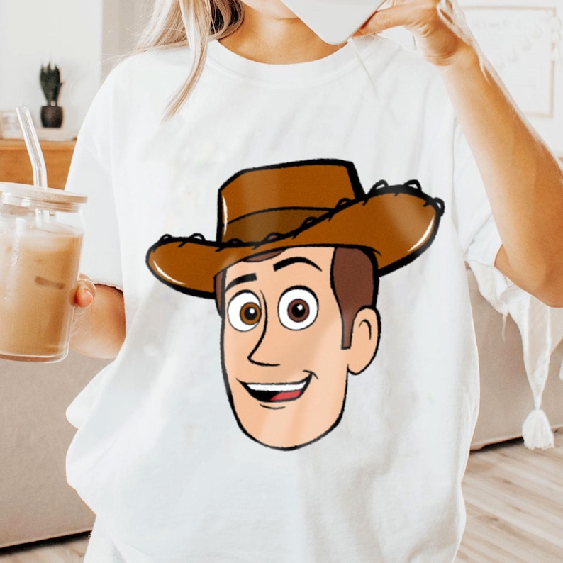 May include: White t-shirt featuring a cartoon illustration of a character with brown hair, a brown cowboy hat, and a smiling face. The character's eyes are brown, and the shirt is plain. The design is centered on the front of the shirt.