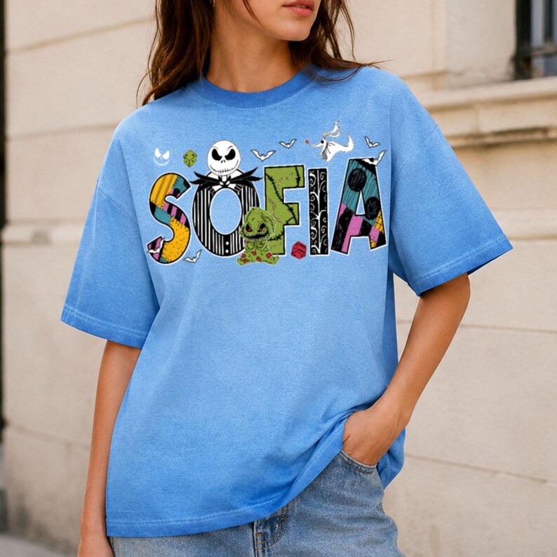 May include: A light blue short-sleeve t-shirt featuring the name SOFIA in large, patterned letters. The design includes characters and motifs from The Nightmare Before Christmas, such as Jack Skellington, Zero, and Oogie Boogie, with bats.