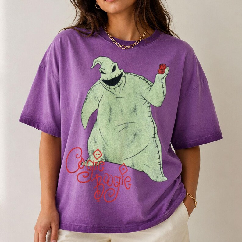 May include: A purple short-sleeve t-shirt featuring a graphic of the character Oogie Boogie from The Nightmare Before Christmas. The character is light green with stitched details, holding two red dice. The text Oogie Boogie is printed in red.