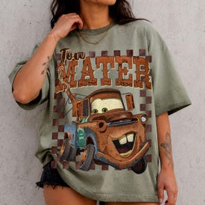 May include: An olive green t-shirt featuring a graphic print of a rusty brown tow truck character with large eyes and a wide smile. Above the truck, the text reads Tow Mater in a distressed brown font, set against a brown and olive checkered background.