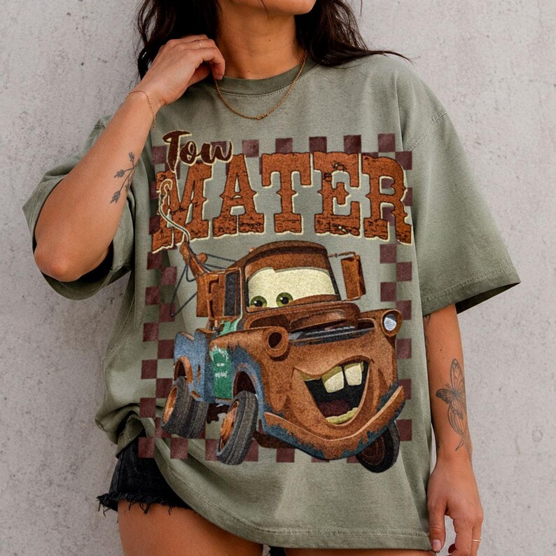 May include: An olive green t-shirt featuring a graphic print of a rusty brown tow truck character with large eyes and a wide smile. Above the truck, the text reads Tow Mater in a distressed brown font, set against a brown and olive checkered background.