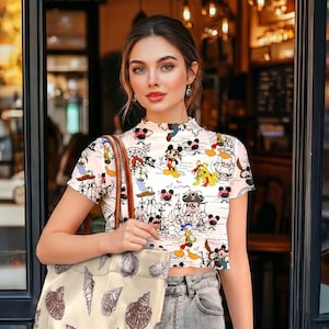May include: A white crop top with a cartoon character print, paired with light wash jeans. A beige tote bag with seashell illustrations is held in the hand. The top features a high neckline and short sleeves.