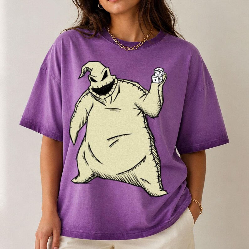 May include: A purple short-sleeve t-shirt featuring a large graphic print of a beige, sack-like character with a wide, jagged-toothed grin. The character is depicted holding two white dice in one hand, with visible black stitching details.