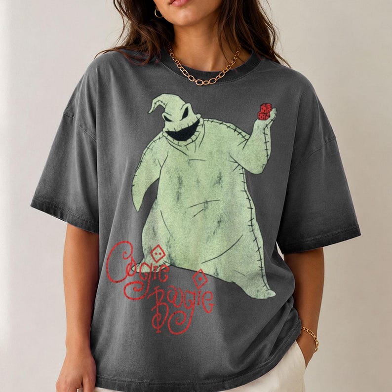 May include: A charcoal gray short-sleeve t-shirt featuring a large graphic of the character Oogie Boogie from The Nightmare Before Christmas. The character is light green, stitched, and holding red dice. The text Oogie Boogie is in red script.