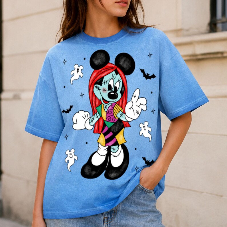 May include: A light blue t-shirt featuring a graphic of a character resembling Minnie Mouse with red hair, stitched skin, and a patchwork dress. The design includes white ghost figures, black bats, and small star accents surrounding the character.