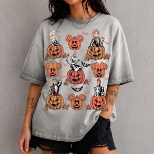 以下が含まれることがあります： ハロウィーンをテーマにしたグラフィックプリントが施されたグレーのアシッドウォッシュTシャツ。スケルトンの衣装を着た様々なキャラクターが、ミッキーマウスの耳の形をしたオレンジ色のジャック・オー・ランタンの中や隣に座っているデザインです。
