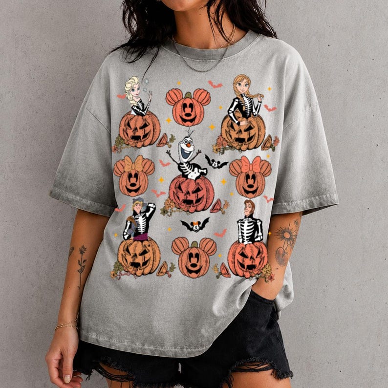 以下が含まれることがあります： ハロウィーンをテーマにしたグラフィックプリントが施されたグレーのアシッドウォッシュTシャツ。スケルトンの衣装を着た様々なキャラクターが、ミッキーマウスの耳の形をしたオレンジ色のジャック・オー・ランタンの中や隣に座っているデザインです。