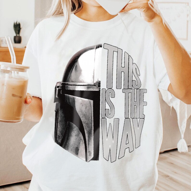 May include: White t-shirt featuring a graphic design of a Mandalorian helmet split in half with the text "THIS IS THE WAY" in gray. The design is centered on the shirt. The shirt is made of a soft material.