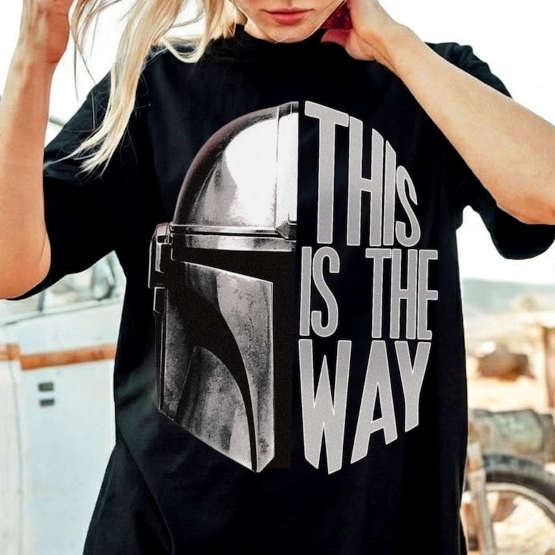 May include: Black t-shirt featuring a graphic of a Mandalorian helmet and the text "THIS IS THE WAY" in white. The design is centered on the front of the shirt. The shirt is made of a soft material.