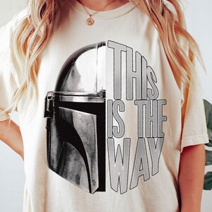 May include: Cream-colored t-shirt featuring a black and gray graphic of a helmet and the text "THIS IS THE WAY" in gray. The design is centered on the front of the shirt. The shirt has short sleeves and a crew neck.