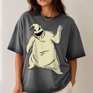 May include: A charcoal gray short-sleeve t-shirt featuring a large graphic print of a beige, sack-like character with a stitched appearance, a wide jagged mouth, and pointed head, holding two white dice in one hand against a dark background.