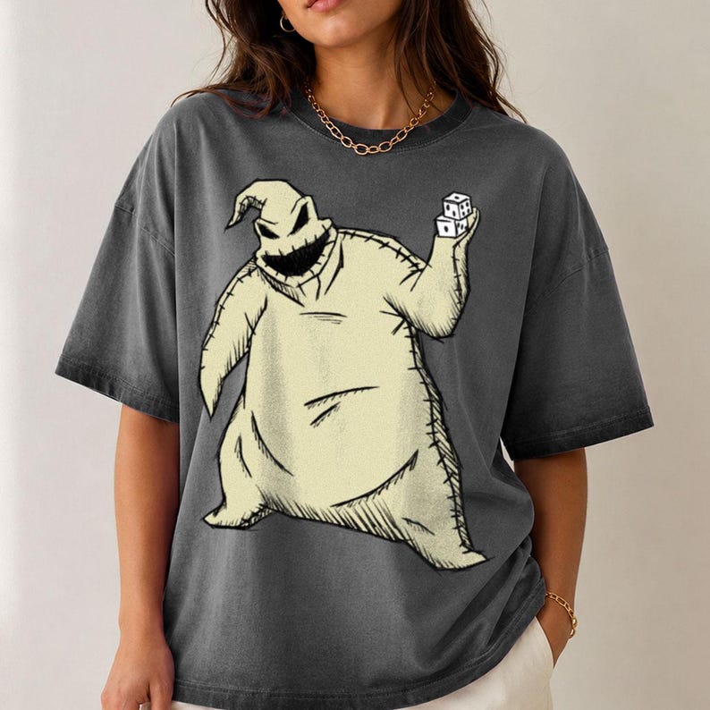 May include: A charcoal gray short-sleeve t-shirt featuring a large graphic print of a beige, sack-like character with a stitched appearance, a wide jagged mouth, and pointed head, holding two white dice in one hand against a dark background.