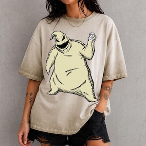 May include: A beige, acid-washed short-sleeve t-shirt featuring a large, centered graphic illustration of a sack-like character with a wide, stitched grin, holding a pair of white dice in one raised hand, rendered in a dark, sketch-like style.