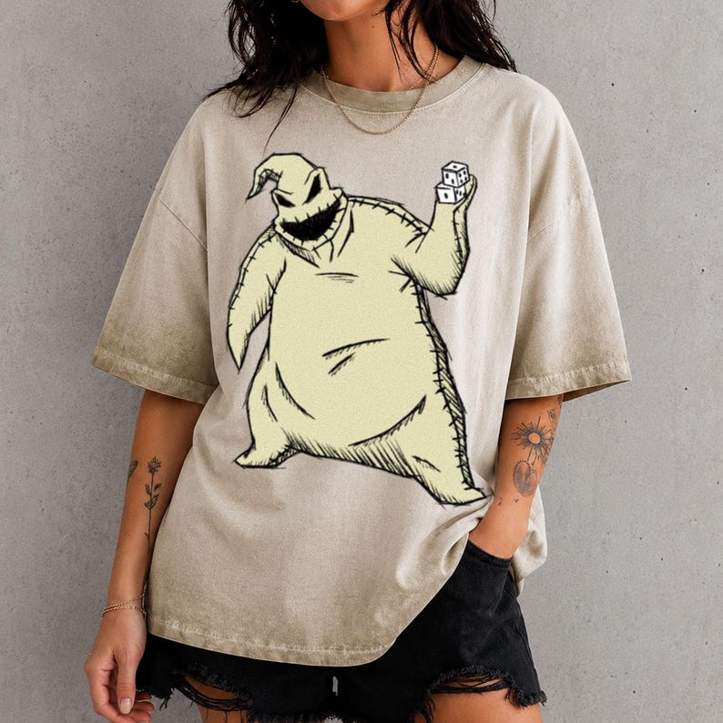 May include: A beige, acid-washed short-sleeve t-shirt featuring a large, centered graphic illustration of a sack-like character with a wide, stitched grin, holding a pair of white dice in one raised hand, rendered in a dark, sketch-like style.