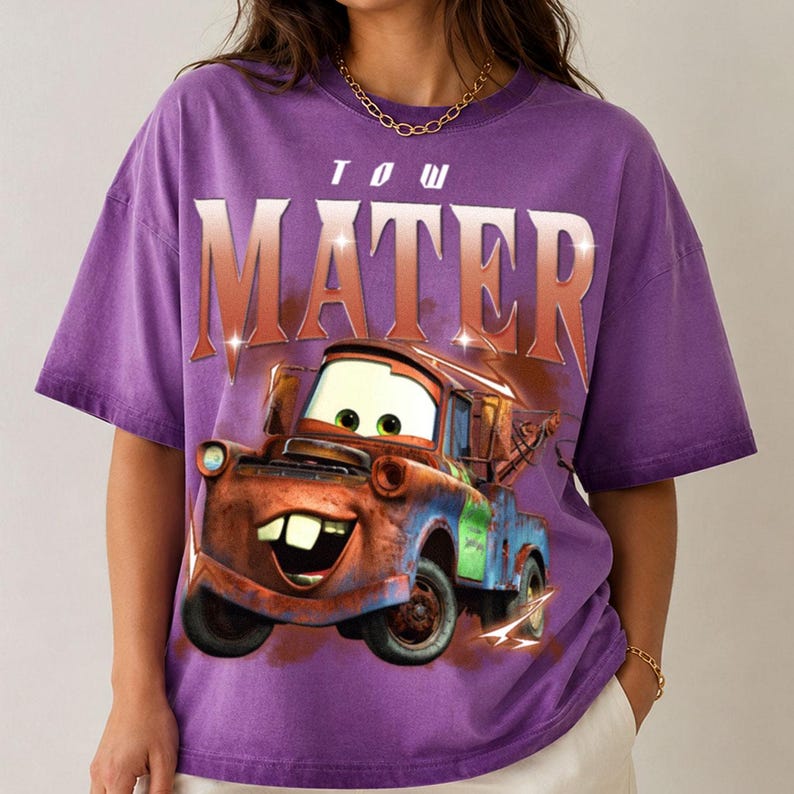K&ouml;nnte beinhalten: Ein violettes Kurzarm-T-Shirt mit einer Grafik der Figur Tow Mater aus dem Film Cars. Der Text TOW MATER ist in gro&szlig;en, fetten Buchstaben mit Farbverlauf &uuml;ber dem rostbraunen Abschleppwagen mit gr&uuml;nen Akzenten auf das T-Shirt gedruckt.