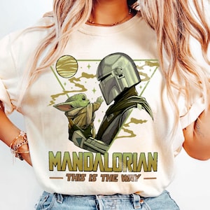 May include: Cream-colored t-shirt featuring a graphic of the Mandalorian and Grogu. The design includes a helmeted figure and a small green creature, with the text "MANDALORIAN - THIS IS THE WAY" in green and gold.