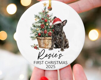 May include: A white ceramic Christmas ornament featuring a watercolor illustration of a French Bulldog wearing a Santa hat next to a decorated Christmas tree. The ornament has the text "Rosie's FIRST CHRISTMAS 2025". A gold string is attached for hanging.