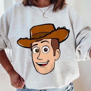 May include: A light gray sweatshirt featuring a cartoon illustration of a smiling cowboy character with a brown hat and hair, and brown eyes. The character's face is centered on the front of the sweatshirt.