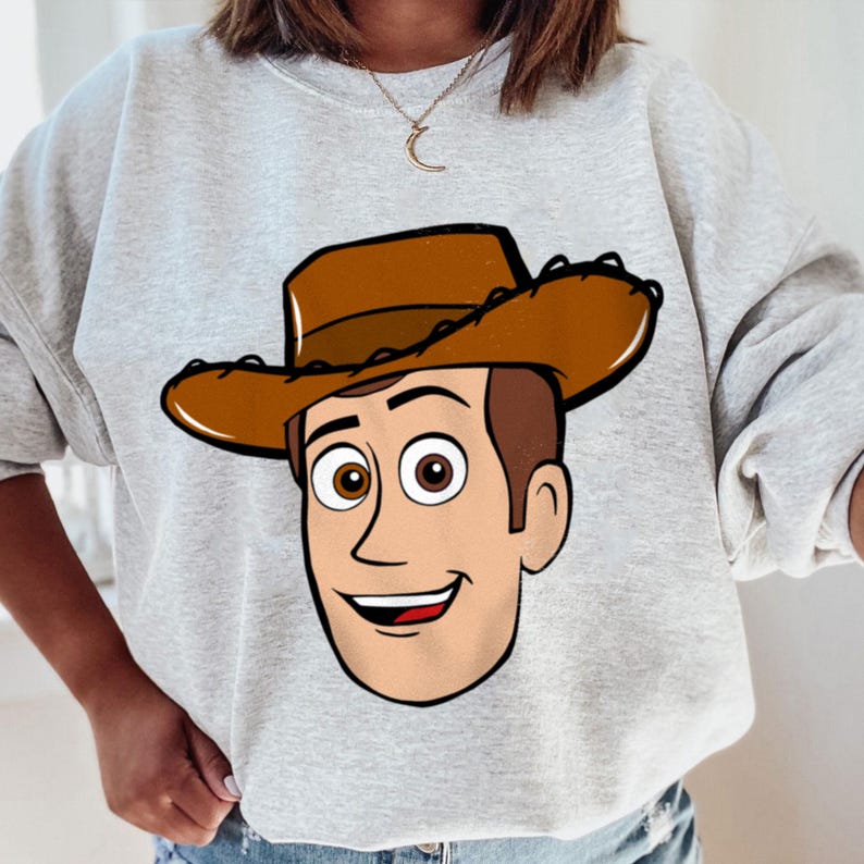May include: A light gray sweatshirt featuring a cartoon illustration of a smiling cowboy character with a brown hat and hair, and brown eyes. The character's face is centered on the front of the sweatshirt.