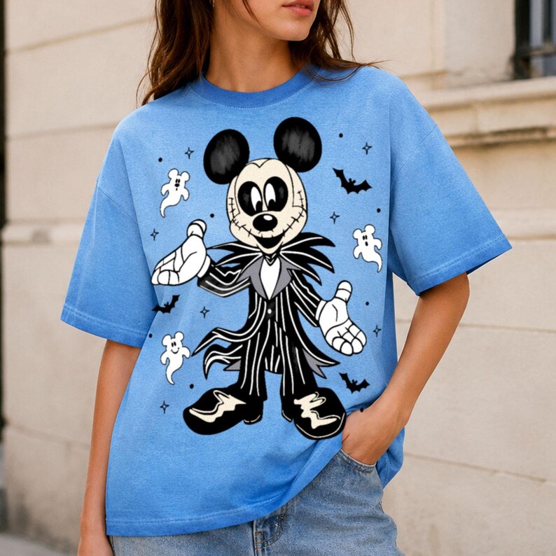 May include: A light blue short-sleeve t-shirt featuring a graphic print of a character with a round head and ears wearing a black and white pinstriped suit. The design includes small white ghost figures, black bats, and four-pointed star accents.