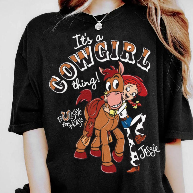 May include: Black t-shirt featuring a cartoon graphic of Jessie from Toy Story hugging Bullseye the horse. The text "It's a Cowgirl thing!" is above the graphic. The shirt is a comfortable, casual style.