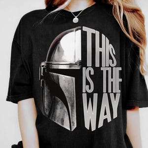 May include: Black t-shirt featuring a silver Mandalorian helmet graphic. The text "THIS IS THE WAY" is printed in white, with the words arranged around the helmet design. The shirt has short sleeves and a crew neck.