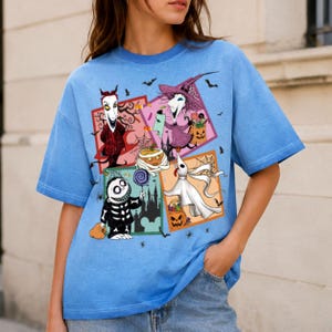 Może przedstawiać: Jasnoniebieski t-shirt z nadrukiem graficznym składającym się z czterech kwadratowych paneli przedstawiających postacie z Miasteczka Halloween, w tym Locka, Shocka, Barrela i Zero, w otoczeniu małych ilustracji nietoperzy i pajęczyn.